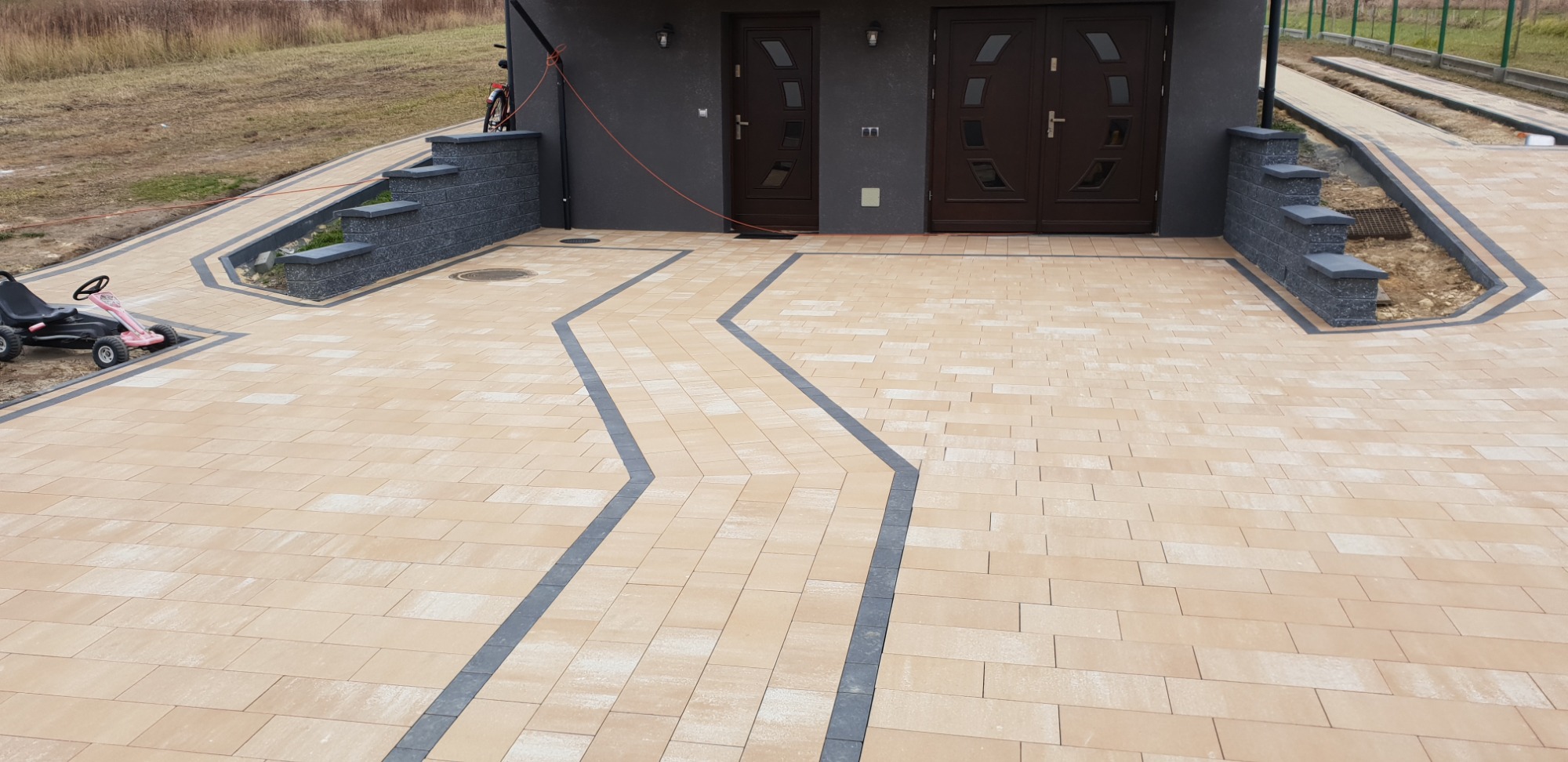 Ułożona kostka brukowa w odcieniach beżu i szarości tworząca wzór zygzaka na podjeździe przed domem z szarą elewacją i brązowymi drzwiami. Widoczny czarny gokart dziecięcy po lewej stronie.
