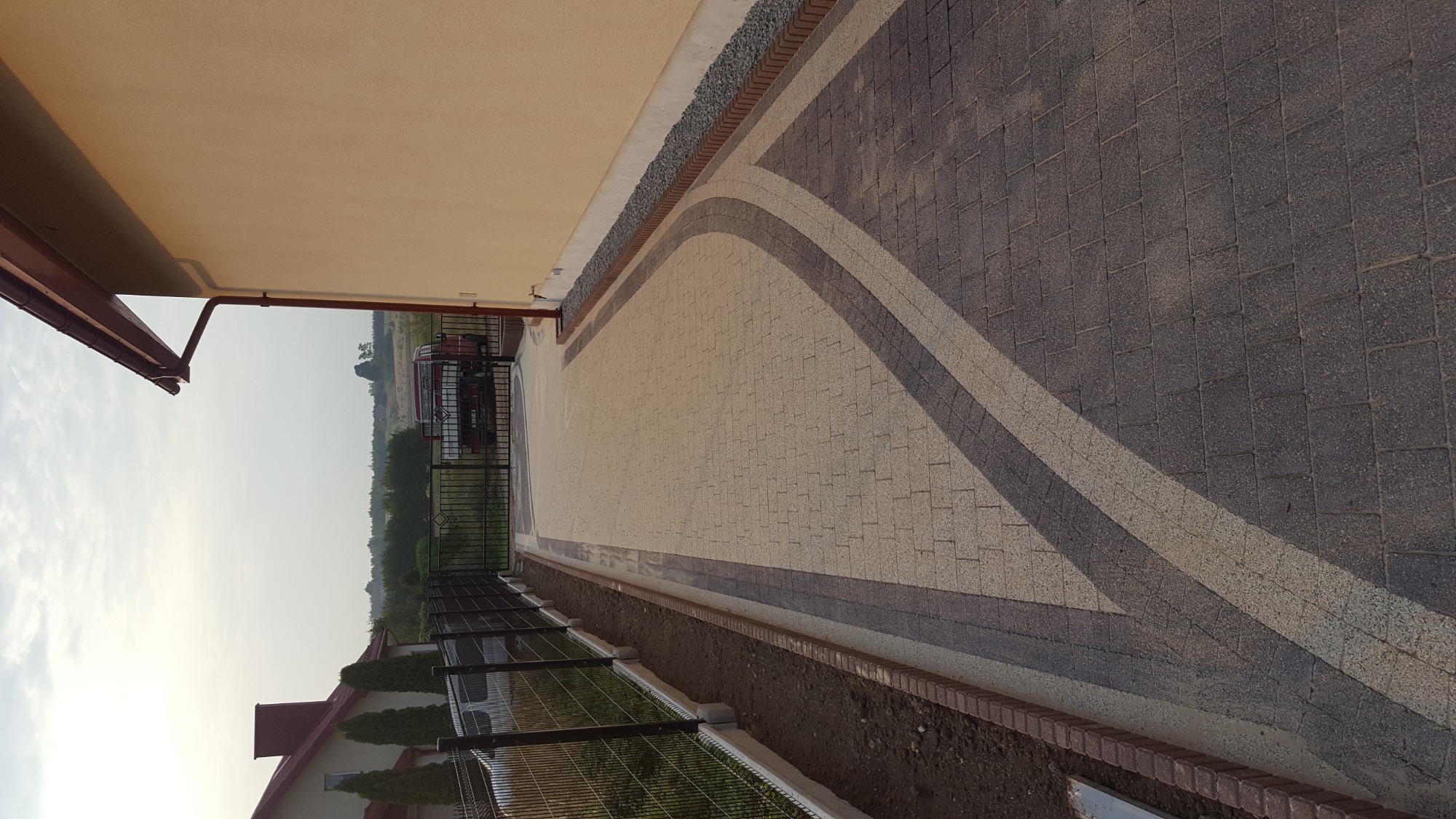 Ułożona kostka brukowa w dwóch odcieniach szarości, tworząca wzór z łukami, widok z góry, obok budynku z szarą elewacją i ogrodzeniem z siatki.