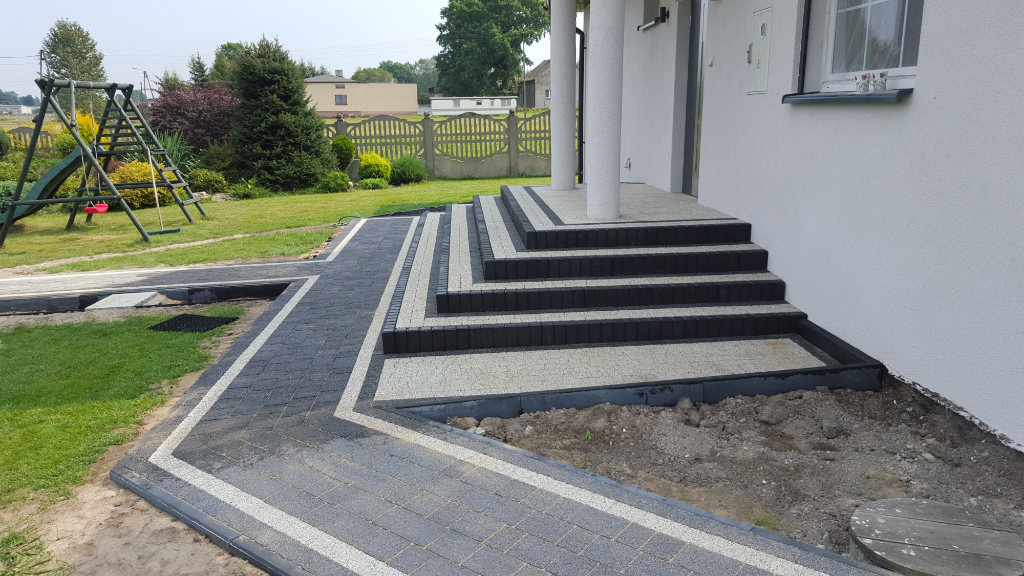 Nowoczesne podejście do domu z kostki brukowej w dwóch odcieniach szarości, z obrzeżami w kontrastującym jasnym kolorze, prowadzące do schodów wejściowych z kolumnami.