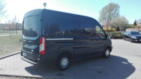 Czarny samochód dostawczy typu van, zaparkowany na parkingu w słoneczny dzień, widoczny z boku, z innym samochodem osobowym w tle.