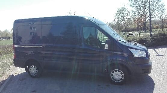 Ciemnoniebieski furgon dostawczy Ford Transit, zaparkowany na szarym brukowanym placu, z budynkiem mieszkalnym i drzewami w tle, uchwycony w słoneczny dzień.