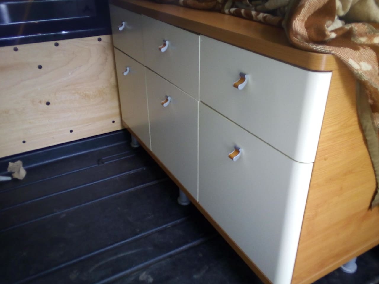 Wnętrze pojazdu z zabudową meblową: komoda z szufladami w jasnych kolorach, zamontowana na podłodze z tworzywa sztucznego, widoczna płyta boczna z otworami montażowymi.