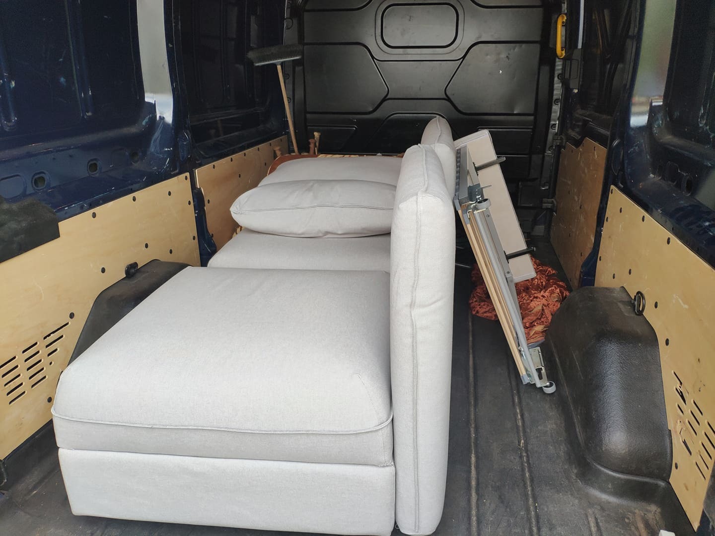 Jasnoszara sofa modułowa z poduszką i składany stelaż łóżka wewnątrz ciemnoniebieskiego busa z drewnianą zabudową ścian.