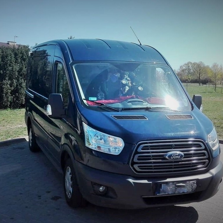 Niebieski bus Ford Transit zaparkowany na brukowanym placu, widok z przodu, antena na dachu, dekoracje na desce rozdzielczej.