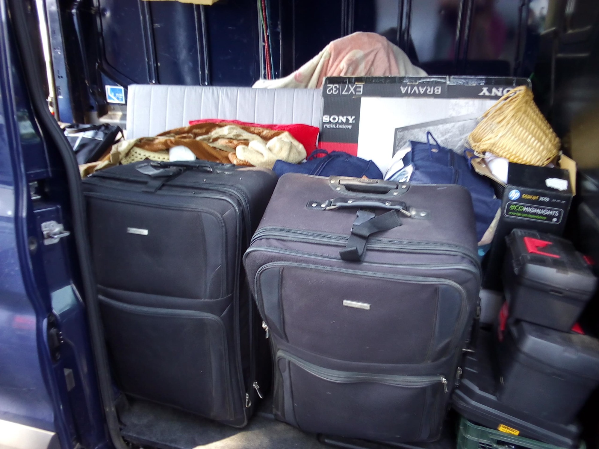 Wnętrze busa załadowane bagażami, kartonami i materacem podczas transportu w Warszawie. Widoczne dwie duże, czarne walizki na kółkach, karton po telewizorze Sony Bravia oraz skrzynki narzędziowe...