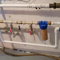 Instalacja wodna z licznymi rurami, zaworami z czerwonymi i niebieskimi uchwytami, filtrem wody w niebieskiej obudowie i z brązowym wkładem oraz niebieskim kluczem do filtra, całość zamontowana...