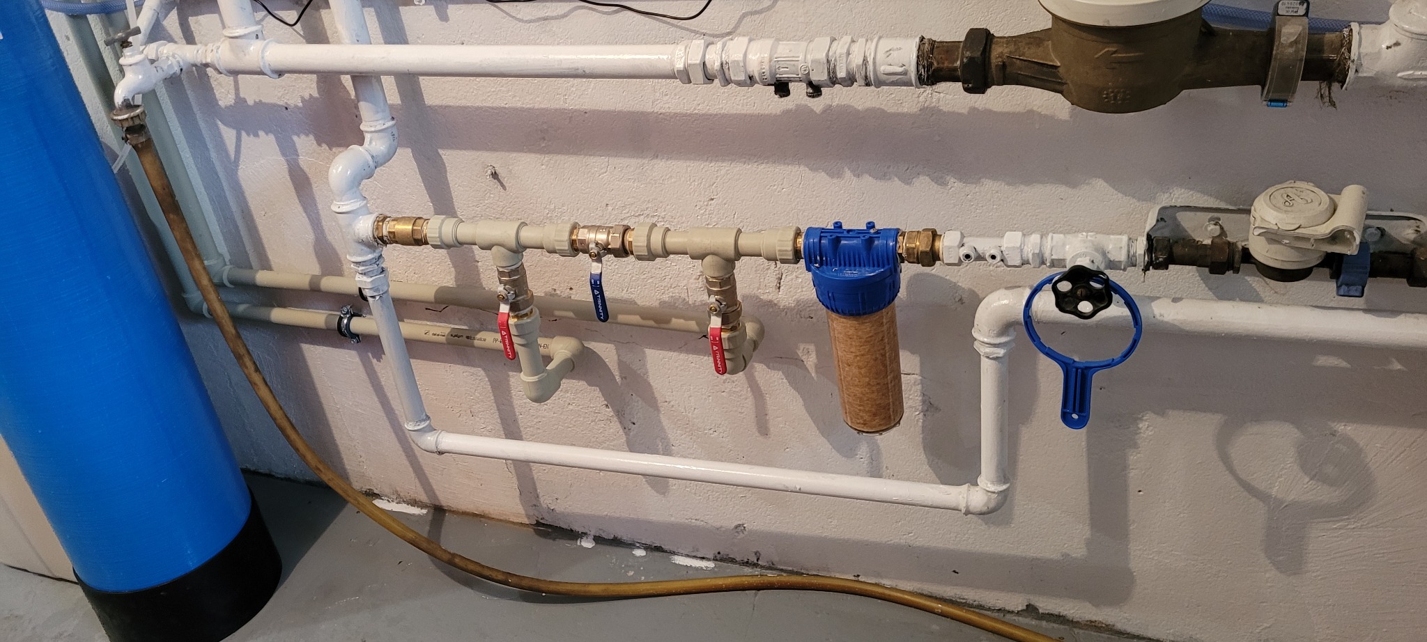 Instalacja wodna z licznymi rurami, zaworami z czerwonymi i niebieskimi uchwytami, filtrem wody w niebieskiej obudowie i z brązowym wkładem oraz niebieskim kluczem do filtra, całość zamontowana...