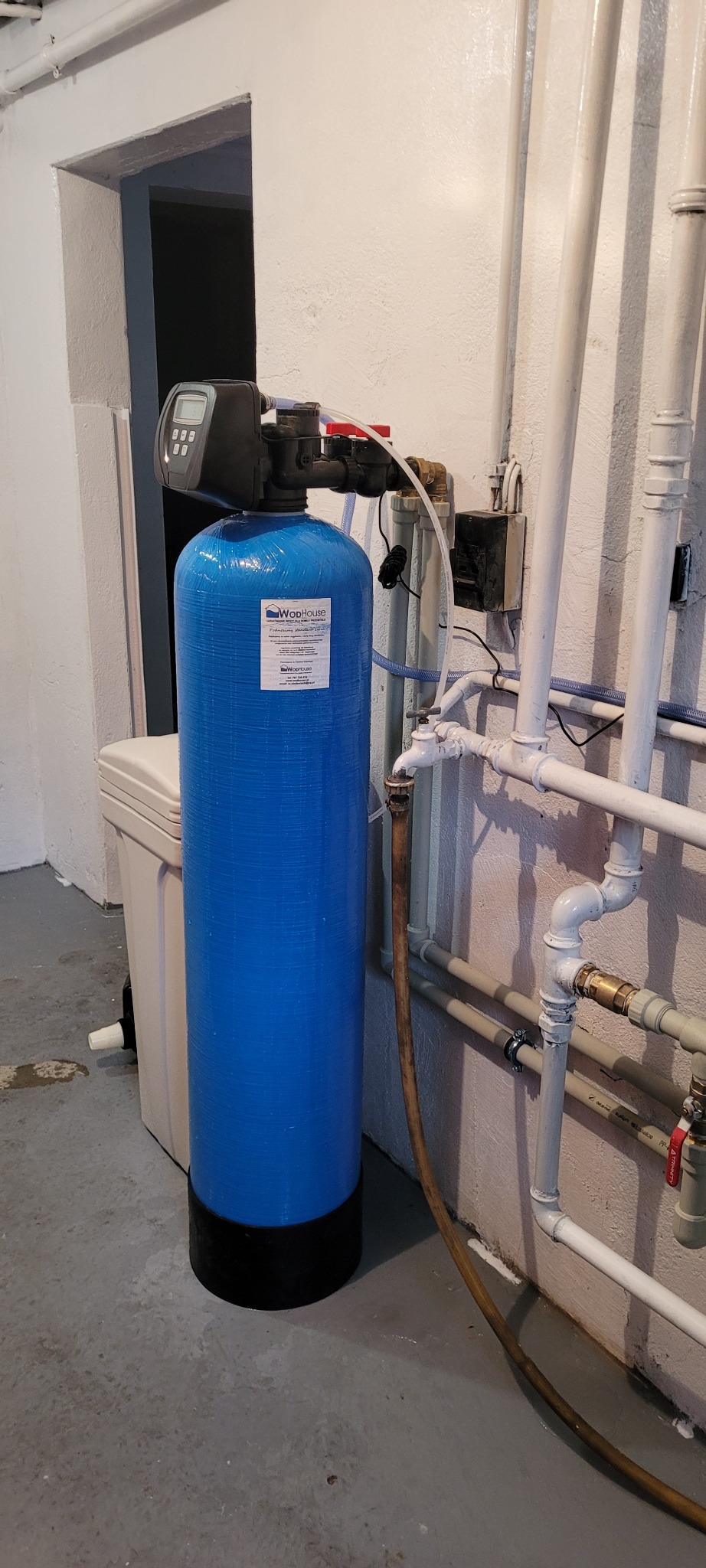 Niebieski filtr wody z elektronicznym sterownikiem, zainstalowany w piwnicy obok białych rur i odpływu.