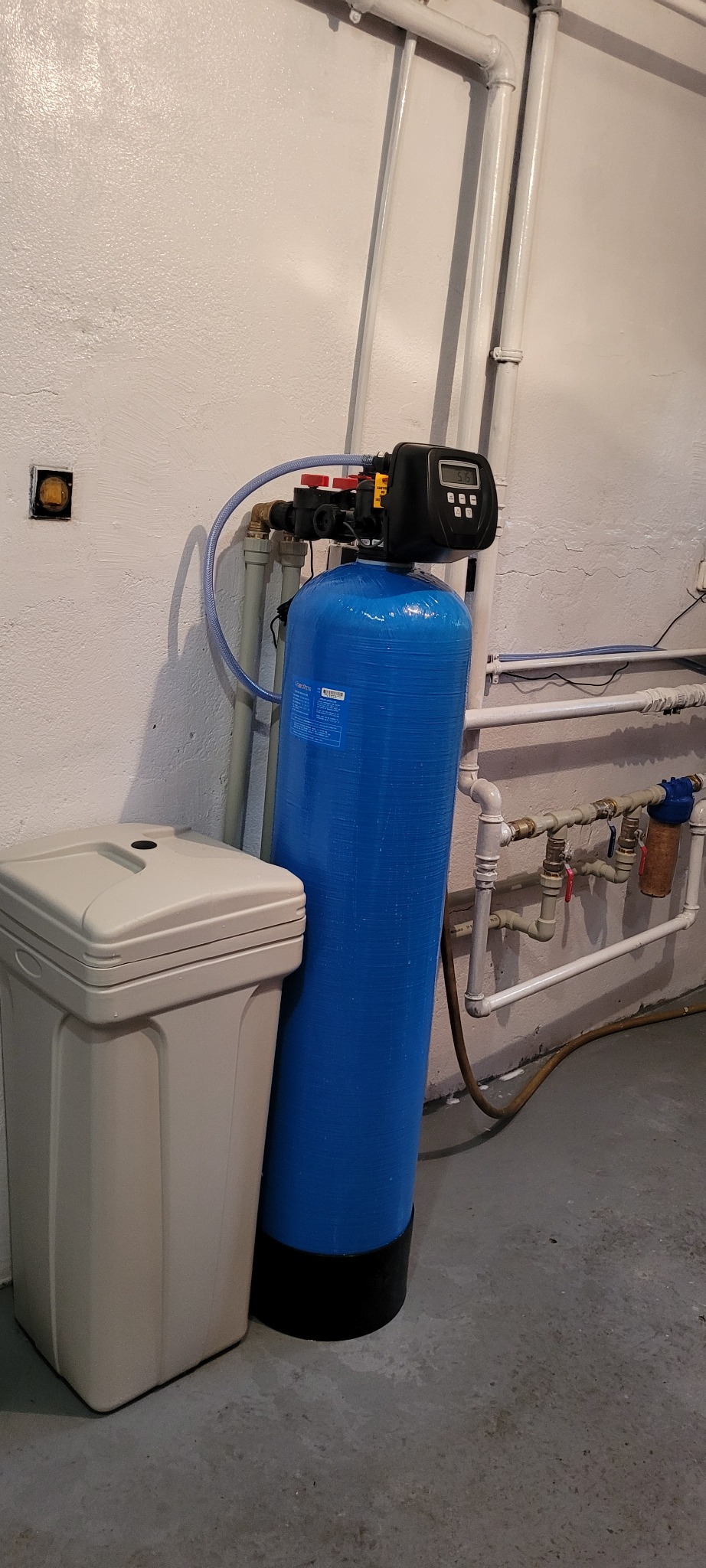 Niebieski zmiękczacz wody z czarnym panelem sterowania i szary pojemnik na sól w piwnicy, widoczne rury instalacji wodnej na ścianie.