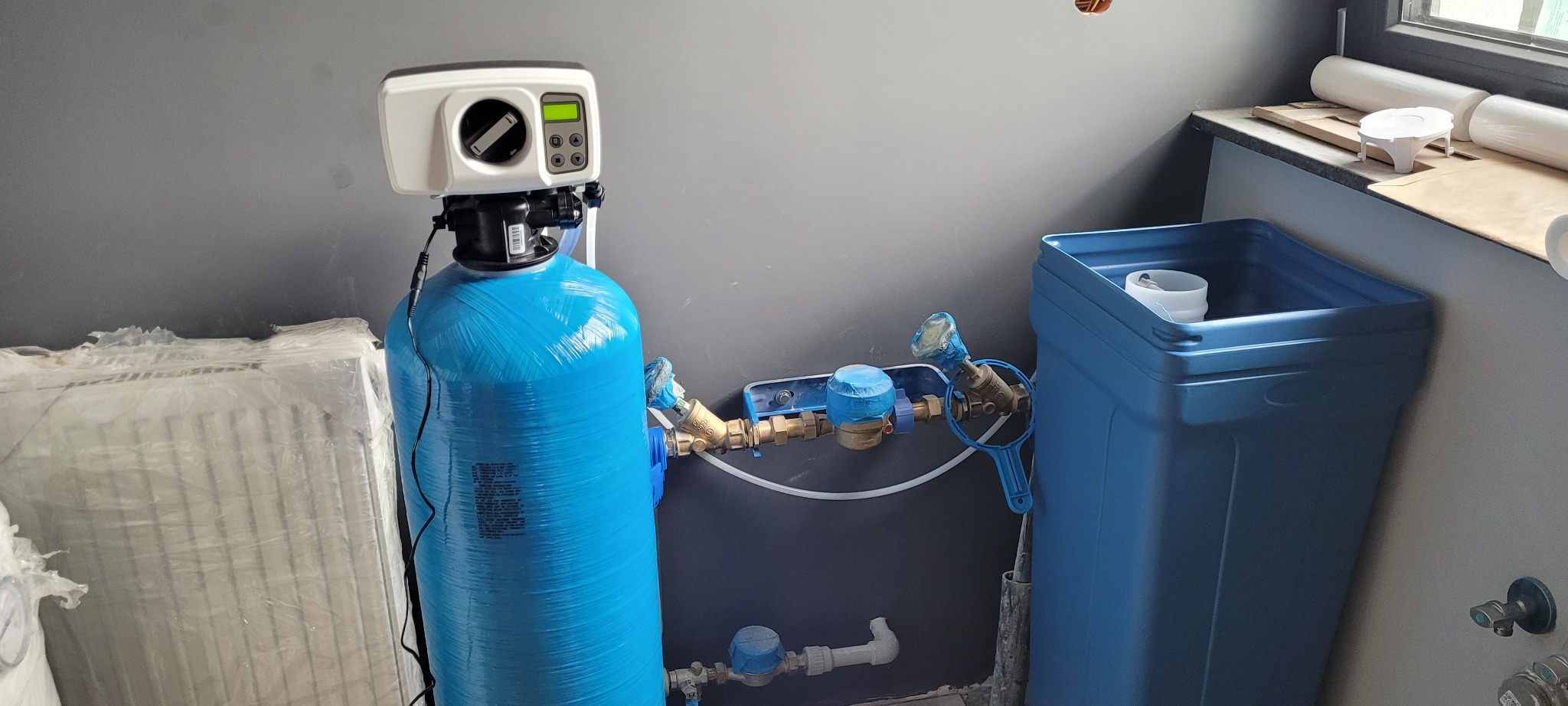 Niebieski zbiornik z elektronicznym sterownikiem i podłączeniami hydraulicznymi, obok niebieski pojemnik, na tle szarej ściany w trakcie prac wykończeniowych.