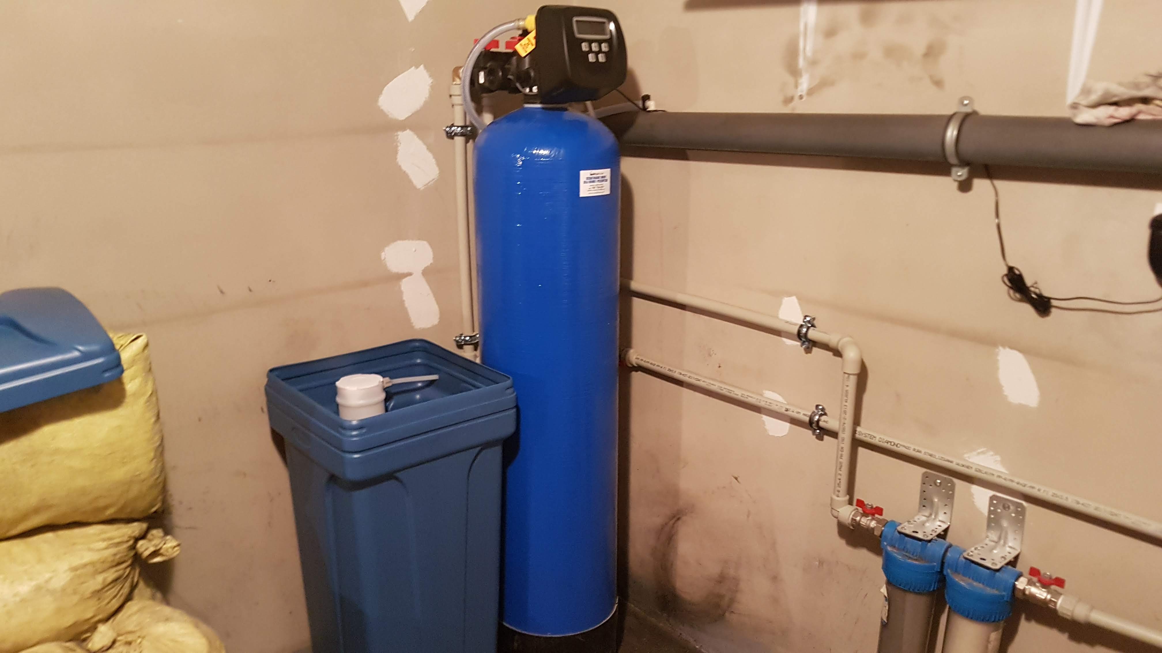 Niebieski system filtracji wody z elektronicznym wyświetlaczem, zbiornik na sól i rury PCV w piwnicy z niedokończonymi ścianami.