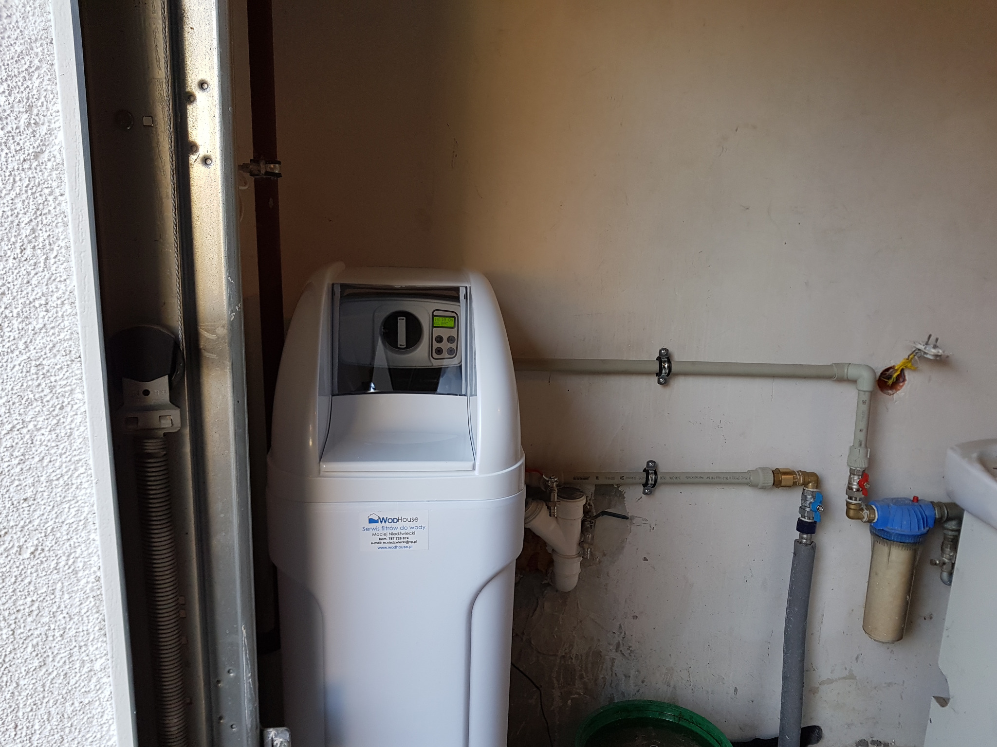 Biały zmiękczacz wody firmy WodHouse z wyświetlaczem LCD, zamontowany w pomieszczeniu gospodarczym z widocznymi rurami instalacji wodnej i filtrem mechanicznym.