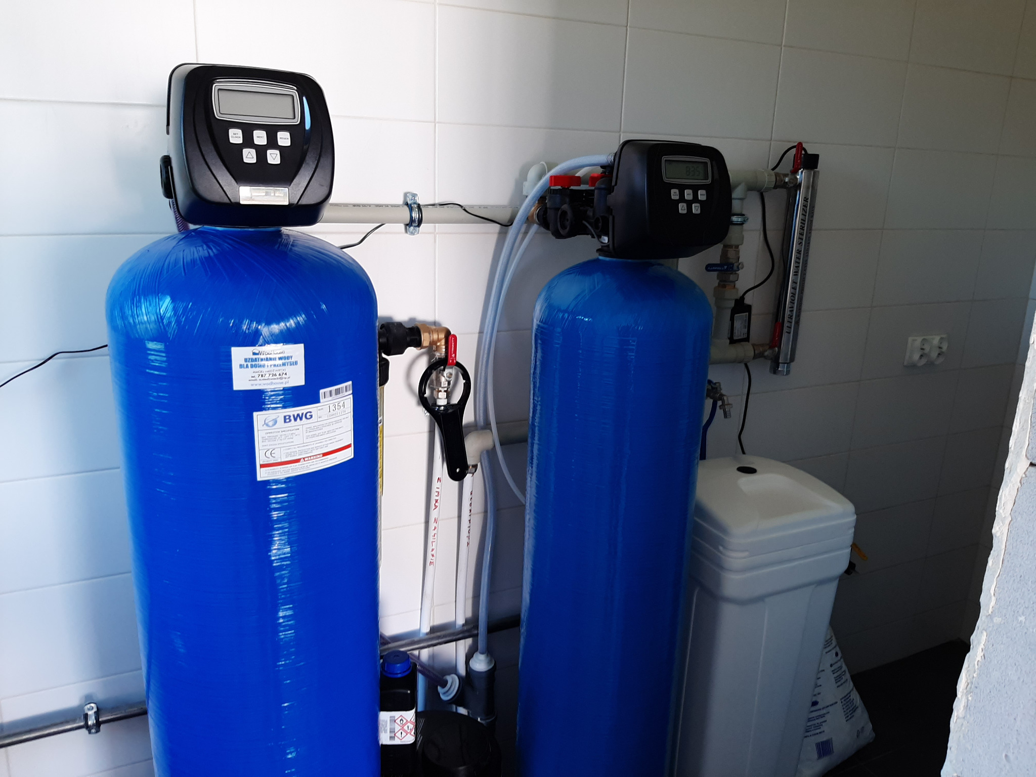 Dwa niebieskie filtry do wody z elektronicznymi panelami sterowania, połączone rurami, obok biały pojemnik na sól. Widoczny również sterylizator wody UV.
