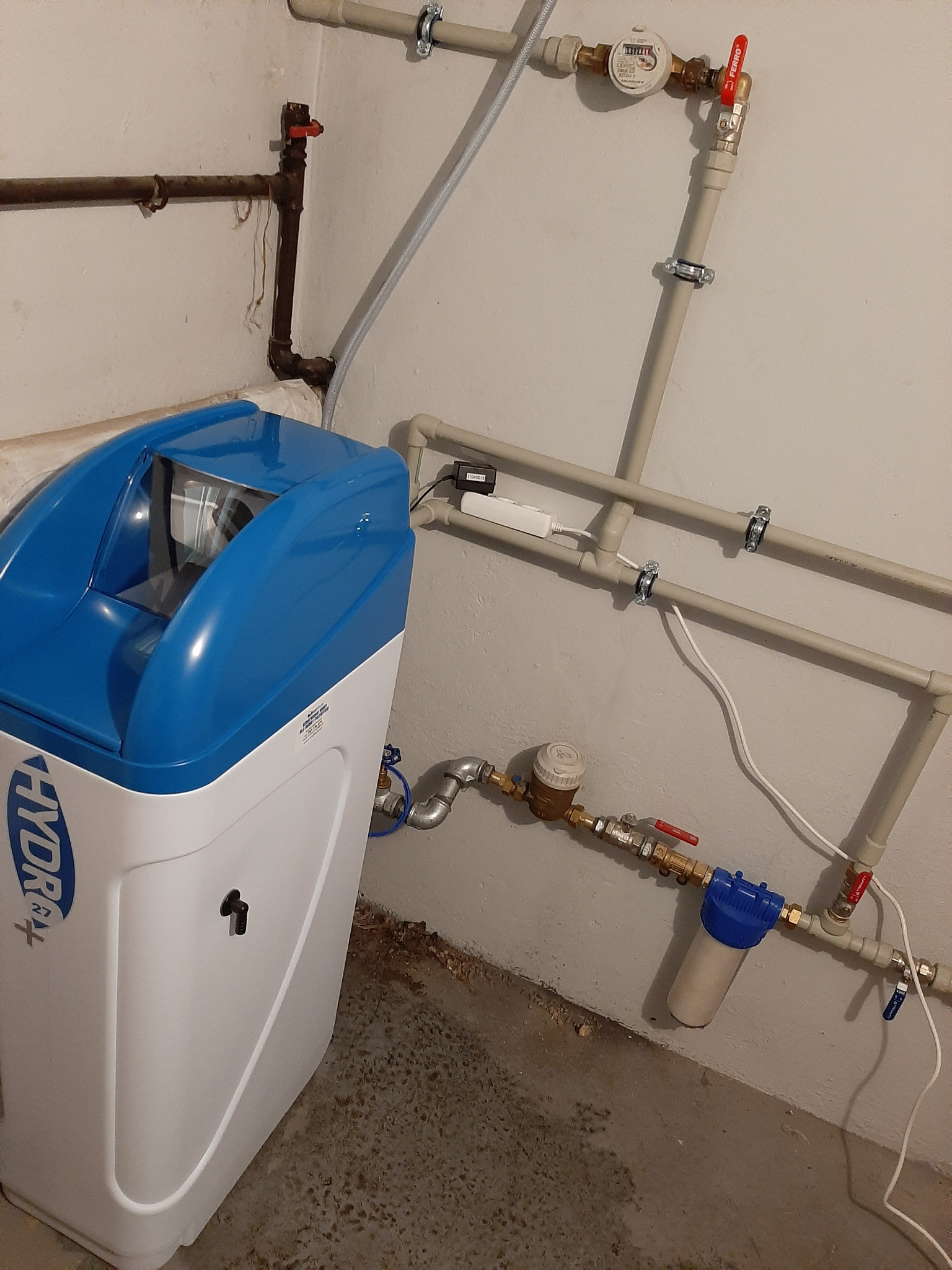 Biało-niebieski zmiękczacz wody z logo Hydropur, podłączony do instalacji wodnej z licznikiem i filtrem, zamontowany w pomieszczeniu technicznym.