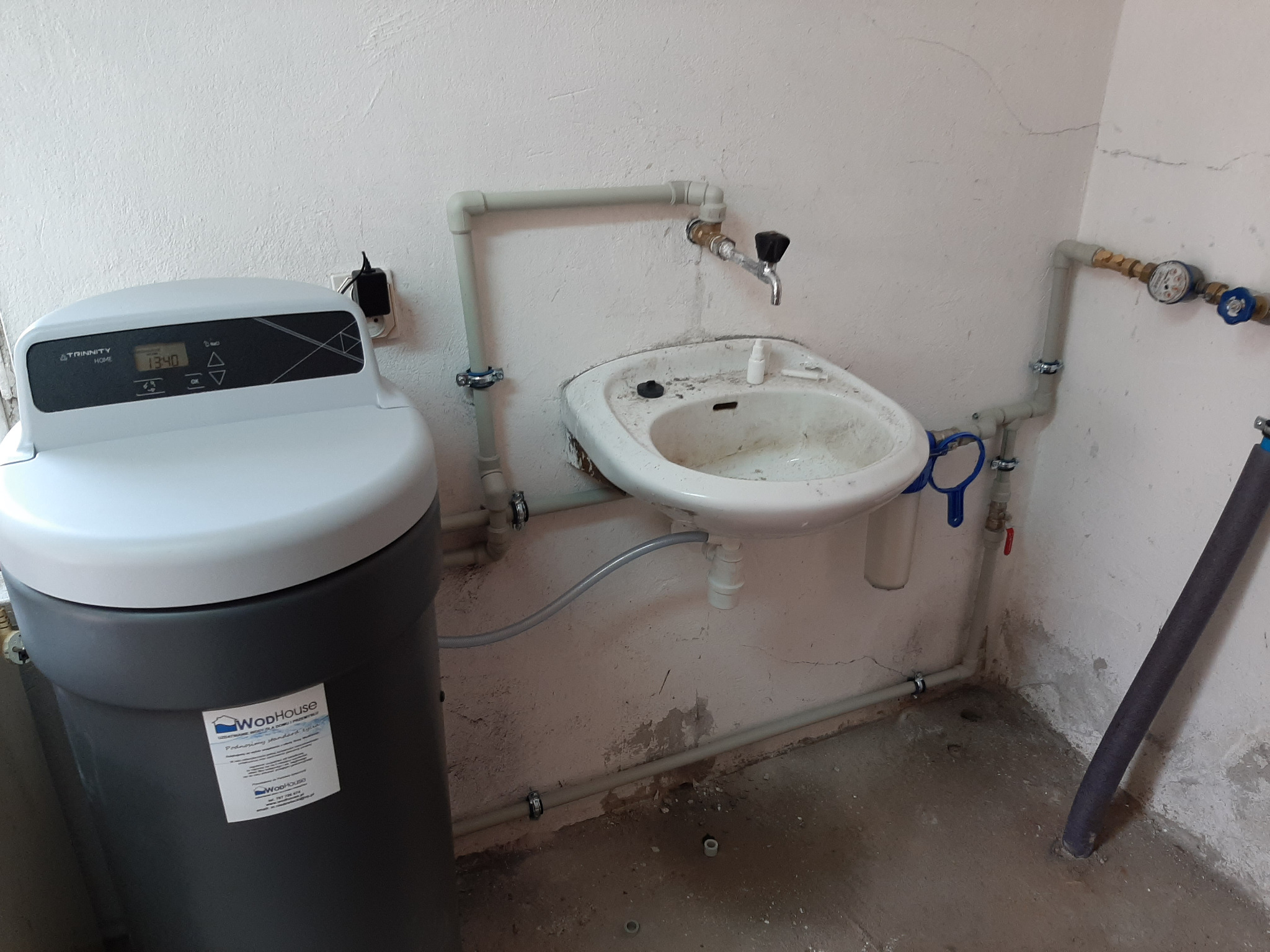 Domowa stacja uzdatniania wody z wyświetlaczem, podłączona do instalacji z umywalką, widoczne rury PCV z zaworami i manometrem, ściany z odpryskami farby.