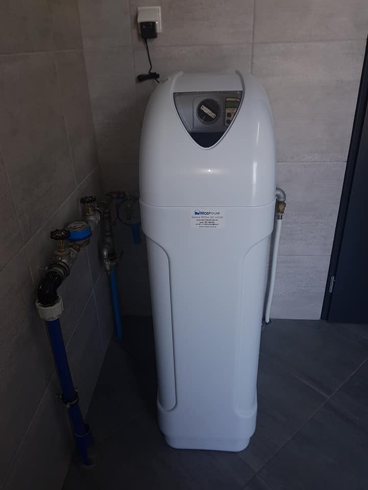 Biały zmiękczacz wody marki WooHouse zainstalowany w rogu pomieszczenia z szarymi płytkami, widoczne rury doprowadzające wodę i zawory.