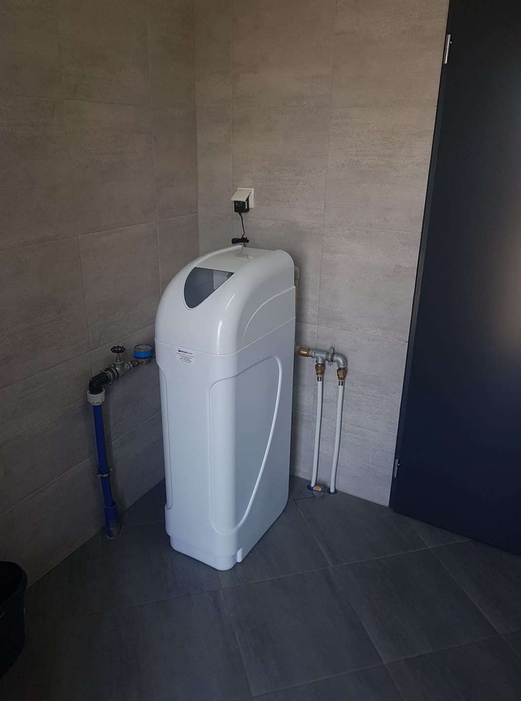 Biały zmiękczacz wody w pomieszczeniu z szarymi płytkami, widoczne podłączenia hydrauliczne i gniazdko elektryczne powyżej urządzenia.