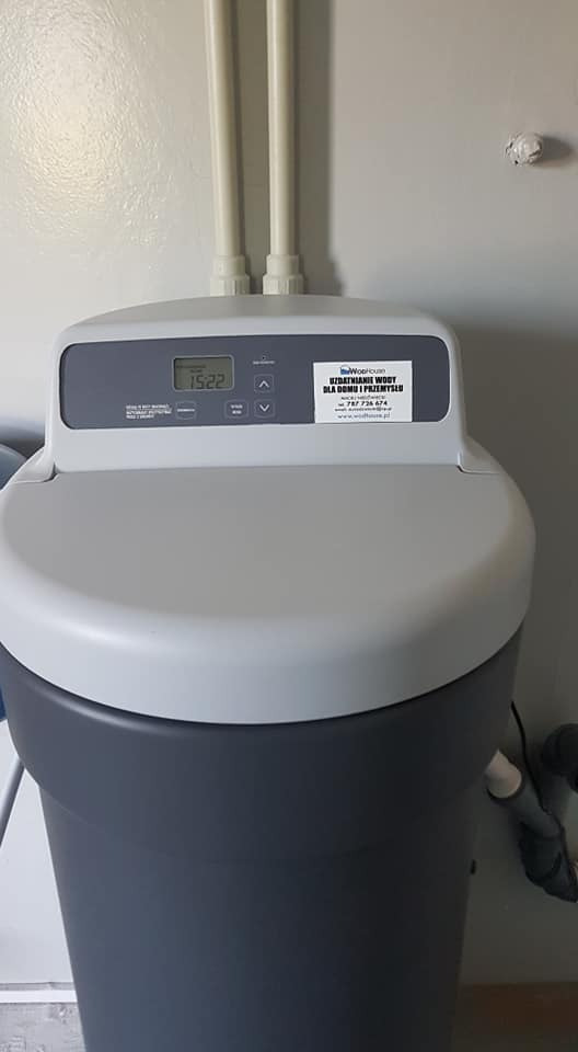 Szaro-białe urządzenie do zmiękczania wody z wyświetlaczem wskazującym godzinę 15:22 i naklejoną etykietą firmy, zainstalowane przy ścianie z rurami.