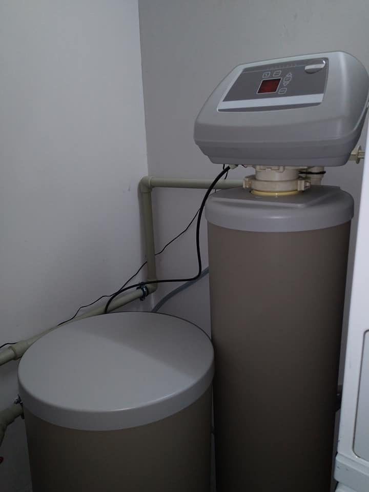 Dwa zbiorniki systemu domowego uzdatniania wody, z szarym sterownikiem elektronicznym na większym zbiorniku, w rogu pomieszczenia z widocznymi rurami instalacyjnymi.