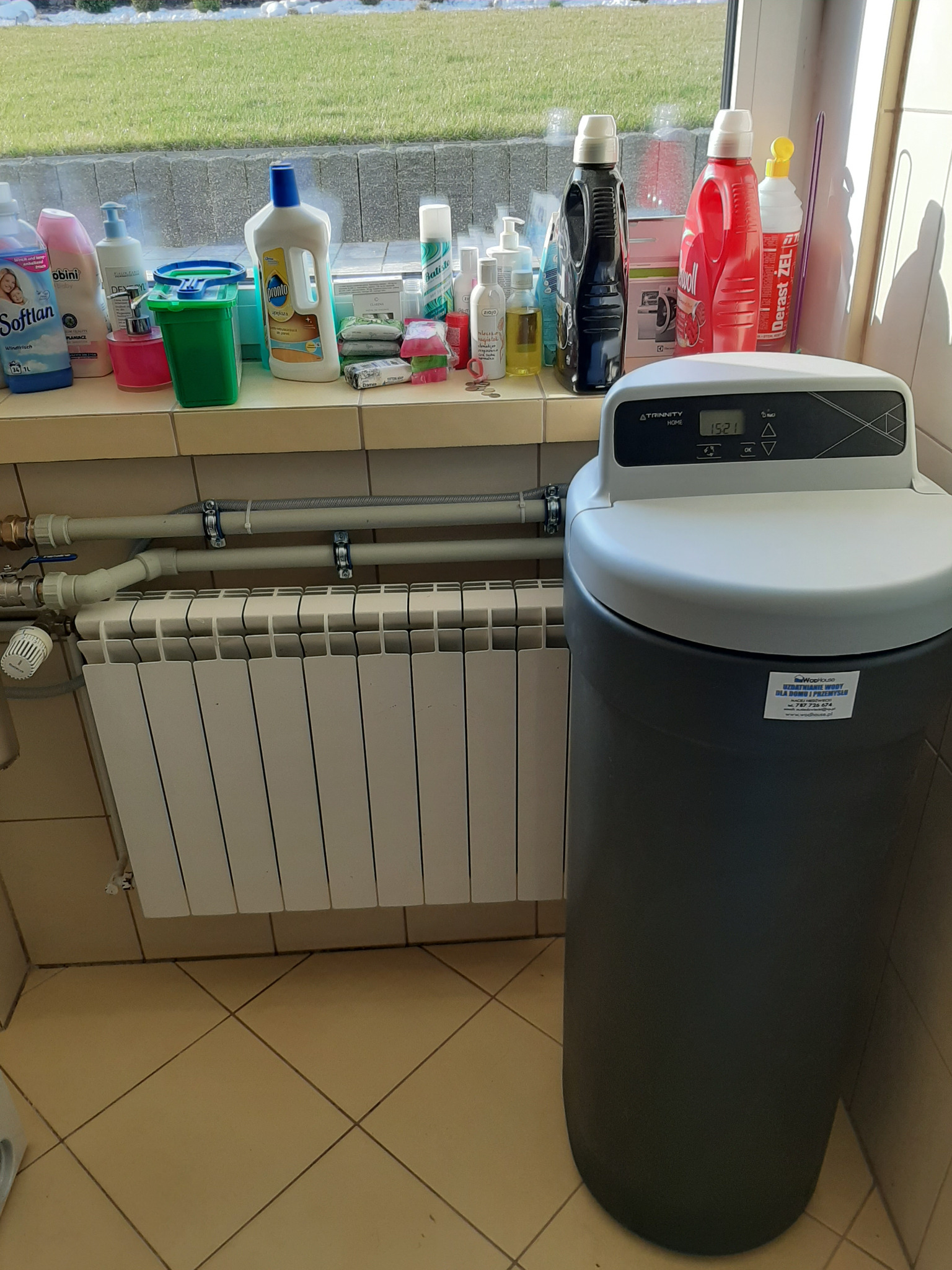 Domowa stacja uzdatniania wody marki Atrinnity Home, ustawiona w pomieszczeniu gospodarczym przy grzejniku i oknie, na parapecie widoczne środki czystości.