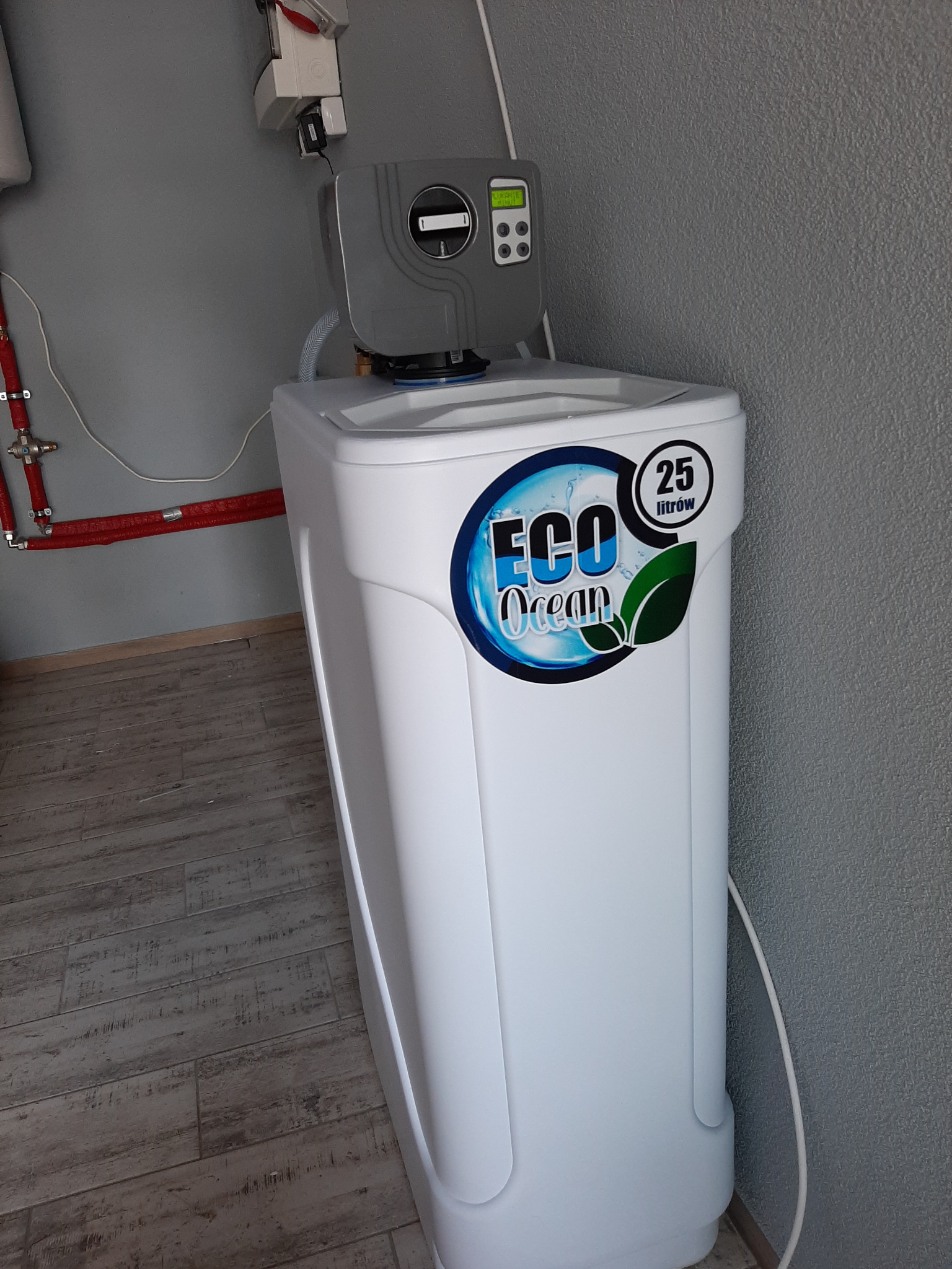 Biały, kompaktowy zmiękczacz wody Eco Ocean 25 litrów, z elektronicznym panelem sterowania, zainstalowany w pomieszczeniu z szarą teksturowaną ścianą i widocznymi czerwonymi rurami instalacji...
