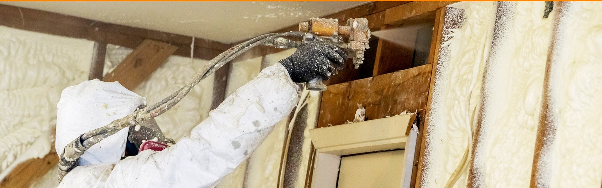 Pracownik w kombinezonie ochronnym aplikuje pianę izolacyjną na drewnianą konstrukcję wewnątrz budynku, widoczne okno z drewnianą ramą.