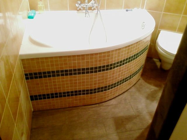 Narożna wanna obudowana mozaiką w odcieniach brązu z ciemnozielonymi pasami, brązowa podłoga imitująca drewno, widoczna toaleta i fragment ściany z brązowymi płytkami.