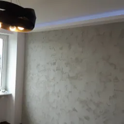 Ściana pokryta dekoracyjną, szarą strukturą, oświetlona od góry listwą LED, z nowoczesną, czarną lampą wiszącą na pierwszym planie i oknem z widokiem na miasto w tle.