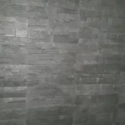 Ściana pokryta szarą okładziną imitującą kamień, ułożoną w poziome pasy z przesunięciem, widoczne łączenia między elementami, oświetlenie z góry.