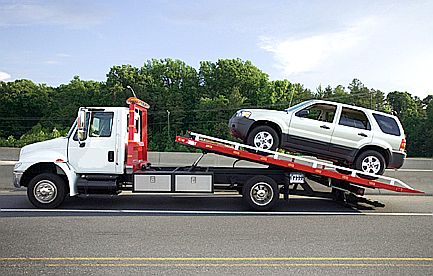 Biała laweta przewozi srebrnego SUV-a na autostradzie w słoneczny dzień, widok z boku, z widocznymi drzewami w tle.
