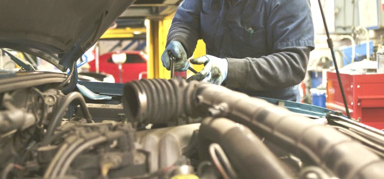 Otwarty silnik samochodowy z widocznymi przewodami i elementami, nad którym pracuje osoba w niebieskim kombinezonie i rękawicach, używając klucza.