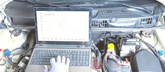 Diagnostyka silnika samochodowego z użyciem laptopa, widoczny wykres parametrów pracy silnika, ręka w rękawicy ochronnej na klawiaturze, wnętrze komory silnika.