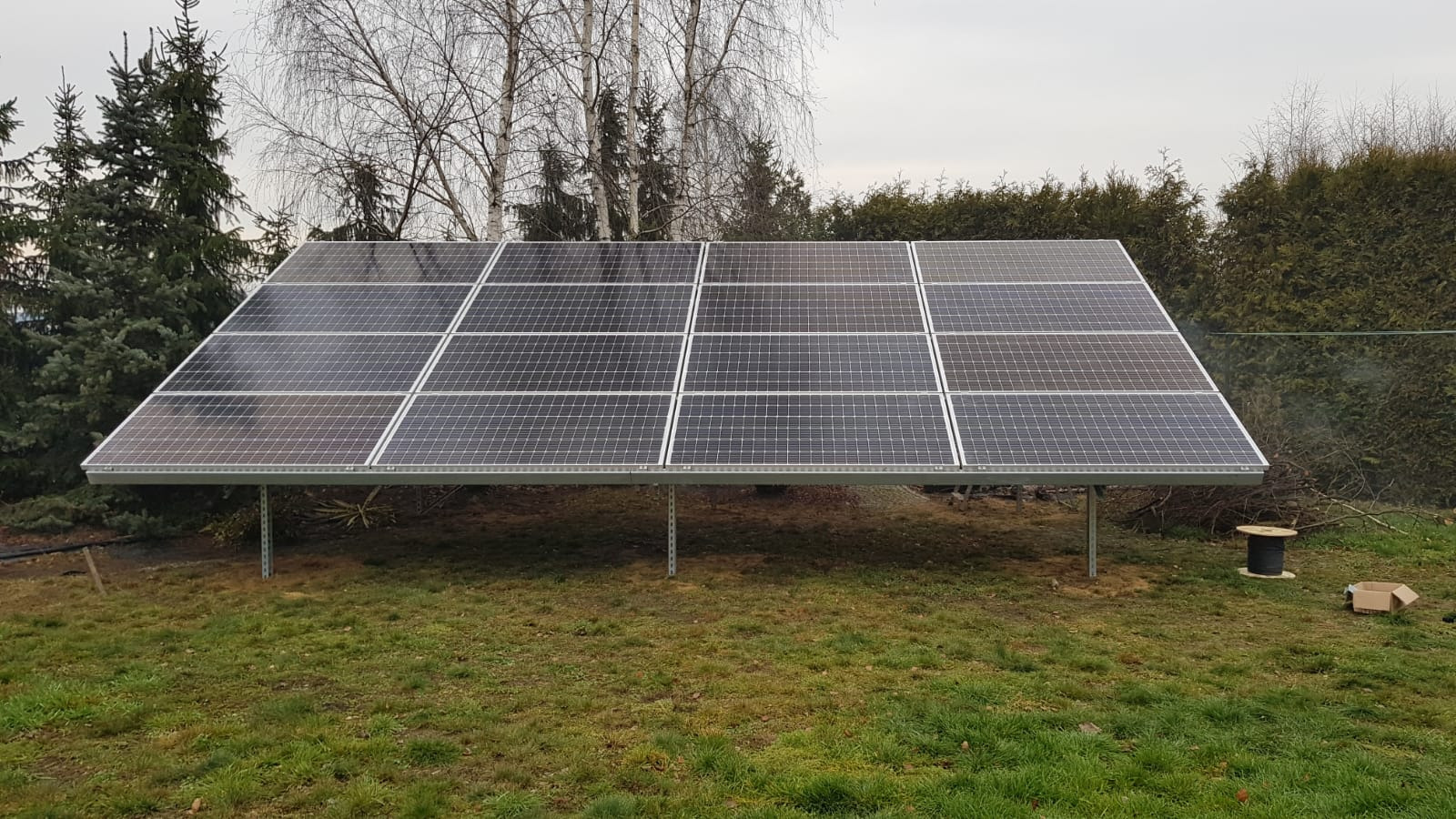 Panel słoneczny na metalowej konstrukcji, ustawiony na trawniku z widoczną szpulą kabla i kartonem w tle, drzewa bez liści i iglaki w tle, pochmurne niebo.