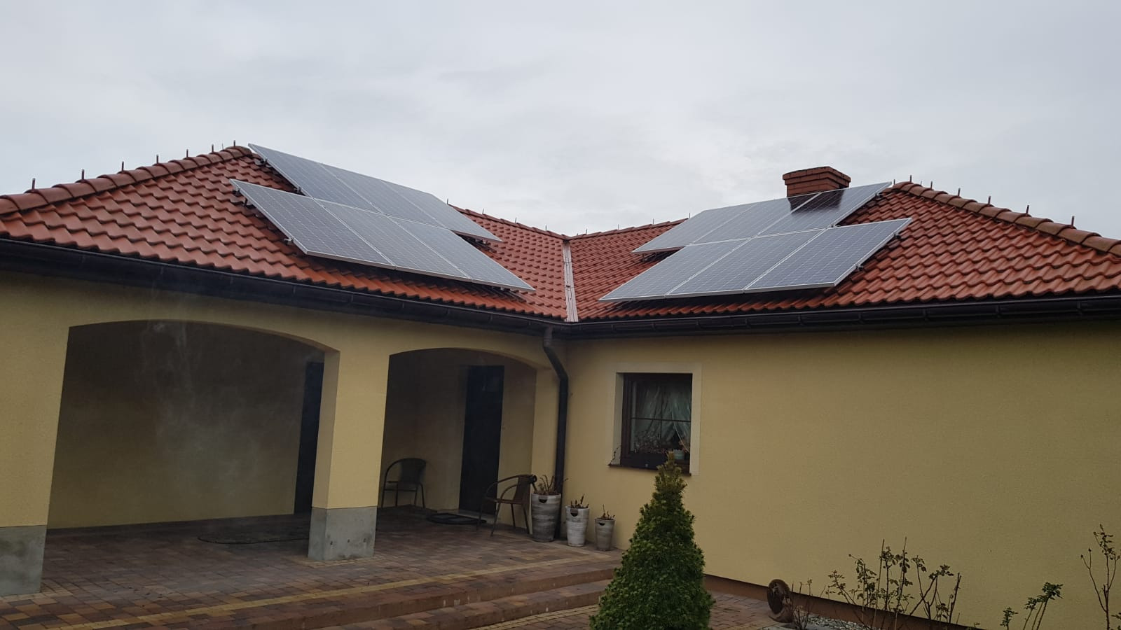 Instalacja fotowoltaiczna na dachu domu z czerwoną dachówką, widok na fragment elewacji z oknem i zadaszonym wejściem.