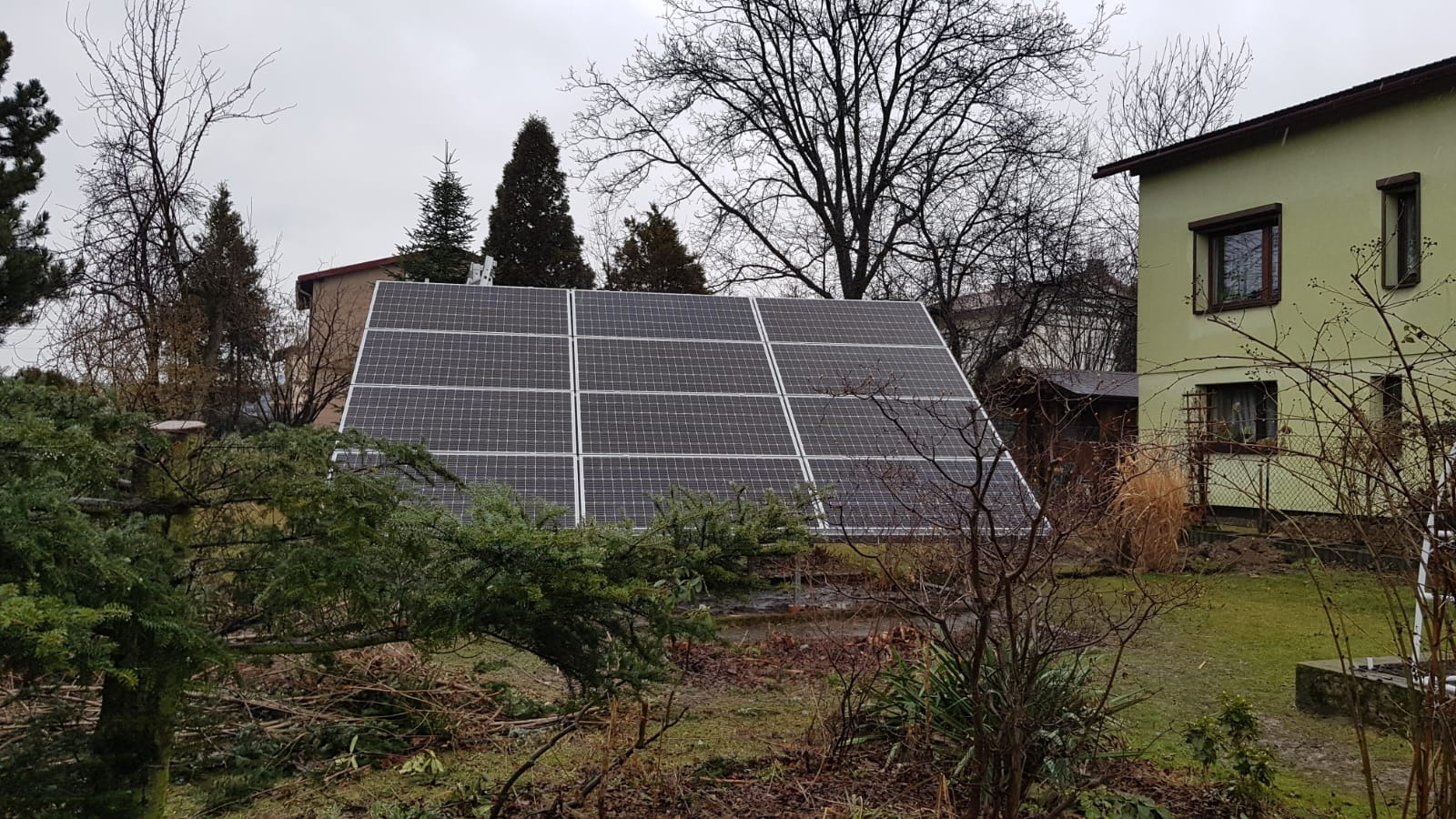 Panel fotowoltaiczny zamontowany na gruncie w Wodzisławiu Śląskim, widoczny zza gałęzi drzew, w tle dom z zieloną elewacją.