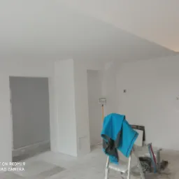 Wnętrze w trakcie malowania na biało, widoczny fragment sufitu z nietypowym załamaniem, ściany przygotowane pod malowanie, drabina z niebieską kurtką roboczą i narzędzia malarskie.