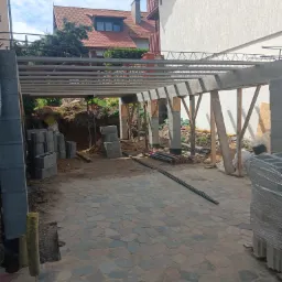 Wznoszenie konstrukcji z betonu i stali na kamiennym podłożu, widoczne bloczki betonowe i elementy drewniane, w tle budynek mieszkalny z czerwonym dachem.