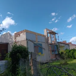 Wznoszenie ścian domu murowanego z betonu komórkowego, widoczne rusztowanie i elementy konstrukcyjne dachu, na pierwszym planie zarośnięte ogrodzenie działki.