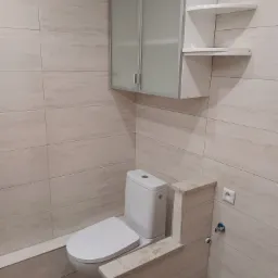 Nowoczesna toaleta z beżowymi płytkami imitującymi kamień, zabudową geberit z tego samego materiału, szafką wiszącą z mlecznymi szybami i narożnymi półkami.