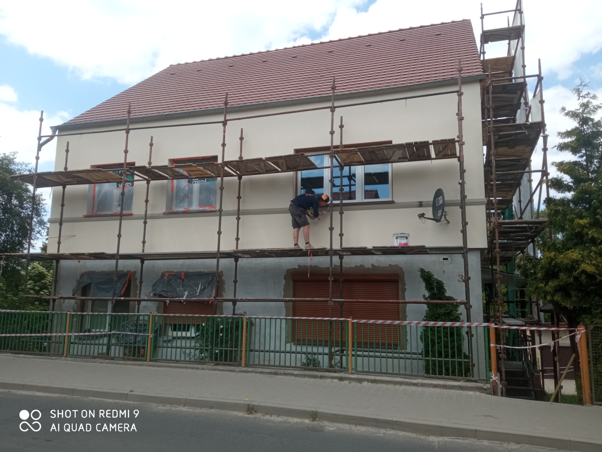 Pracownik na rusztowaniu, szlifuje ścianę domu przed nałożeniem elewacji, widoczny fragment ocieplonej ściany, okna zabezpieczone folią, puszka farby na rusztowaniu.