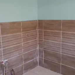 Fragment łazienki w trakcie remontu z brązowymi płytkami imitującymi drewno na ścianach, beżowymi płytkami na podłodze i wystającymi rurami instalacji wodnej