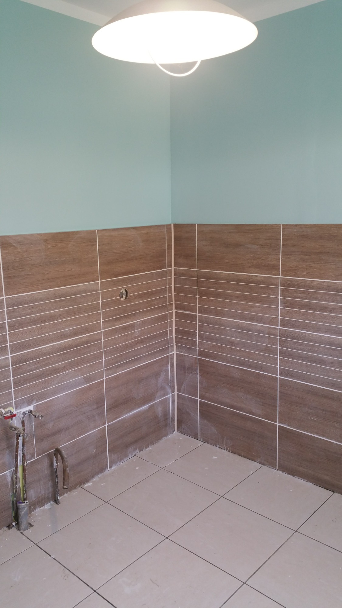 Fragment łazienki w trakcie remontu z brązowymi płytkami imitującymi drewno na ścianach, beżowymi płytkami na podłodze i wystającymi rurami instalacji wodnej