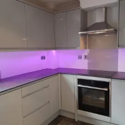 Nowoczesna kuchnia z szarymi, lakierowanymi szafkami, blatem w kolorze ciemnego grafitu i podświetleniem LED w odcieniach fioletu, widoczny piekarnik i okap.