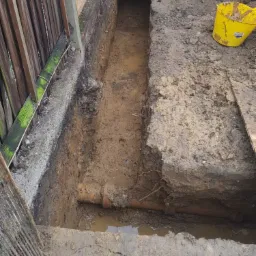 Wykop pod fundament z widoczną rurą kanalizacyjną, częściowo wypełniony wodą, obok znajduje się żółte wiadro budowlane, a w tle widoczny fragment drewnianego płotu.