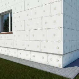 Budynek w trakcie docieplania, widoczne płyty izolacyjne przymocowane do elewacji za pomocą łączników, narożnik budynku z widocznym oknem, trawnik i chodnik przed budynkiem.