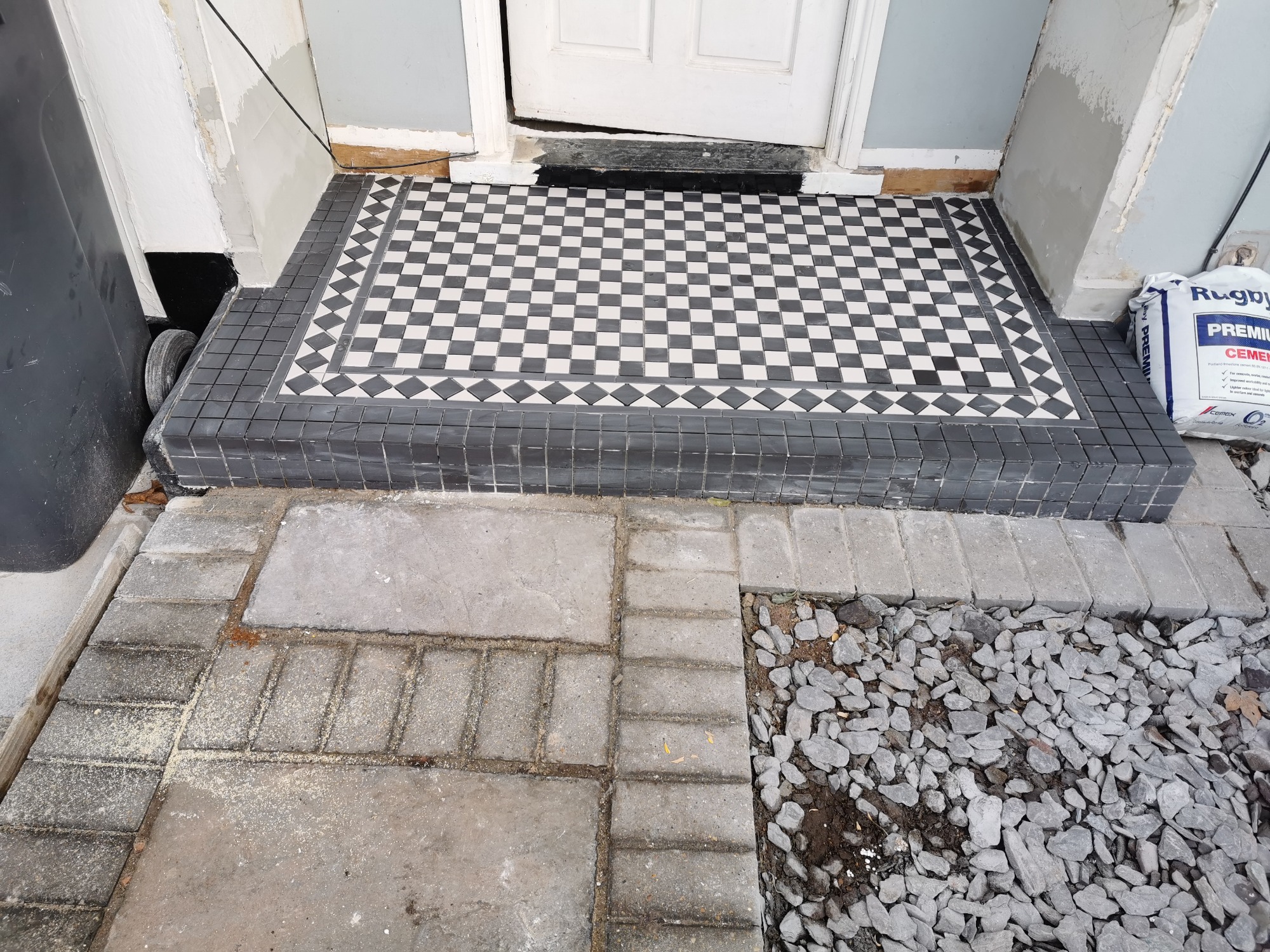 Ułożona czarno-biała mozaika z płytek na schodach wejściowych, widoczny worek cementu i częściowo ułożona kostka brukowa przed wejściem.