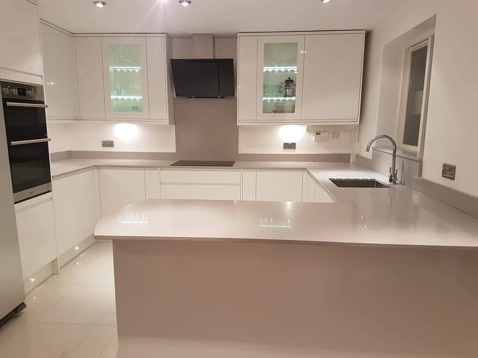 Nowoczesna, jasna kuchnia z białymi, lakierowanymi frontami, szarym blatem i wyspą kuchenną. Oświetlenie LED w szafkach i pod szafkami podkreśla minimalistyczny design. Widoczny zlew z chromowaną...