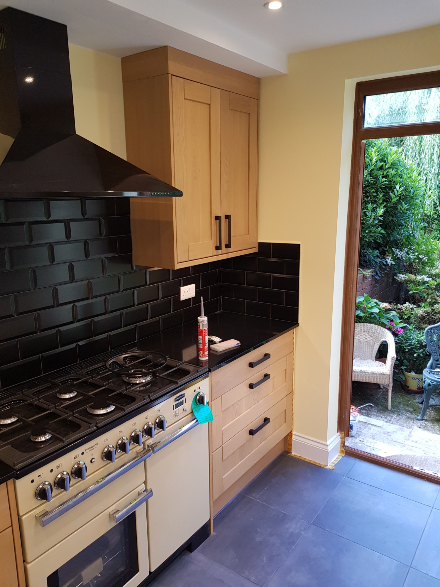 Wykończona kuchnia we Włocławku z czarnymi płytkami metra na ścianie, jasnymi drewnianymi szafkami i dużym beżowym piecem. Widok na zielony ogród przez otwarte drzwi.