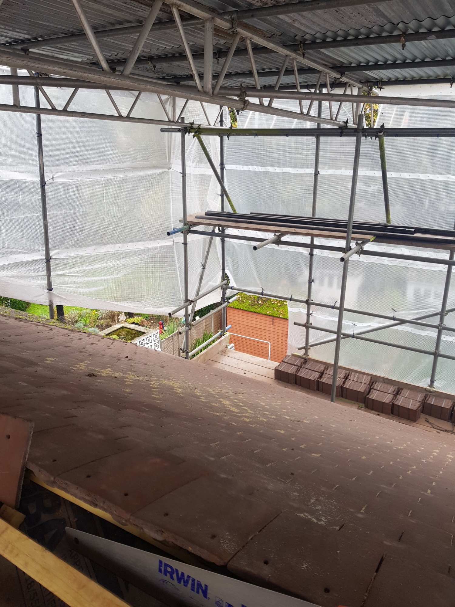 Widok z góry na dach pokryty starą dachówką, przygotowany do remontu, z rozstawionym rusztowaniem i zabezpieczającą folią, w tle widoczny ogród.