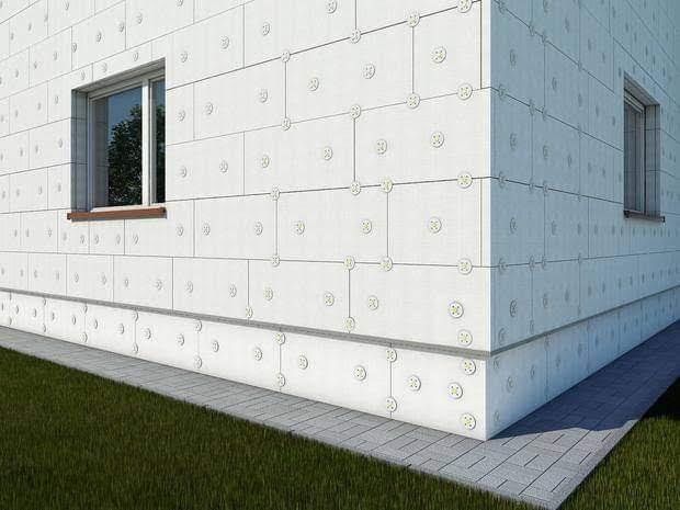 Budynek w trakcie docieplania, widoczne płyty izolacyjne przymocowane do elewacji za pomocą łączników, narożnik budynku z widocznym oknem, trawnik i chodnik przed budynkiem.